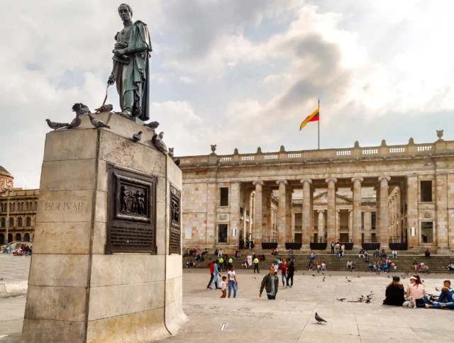 Statue of Simón Bolívar on a pedestal with pigeons. People sit and gather in front of the large neoclassical building, adorned with a Colombian flag under a cloudy sky.