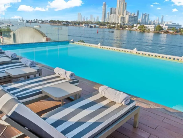 Rooftop infinity pool with striped lounge chairs overlooking a city skyline and waterway under a partly cloudy sky.