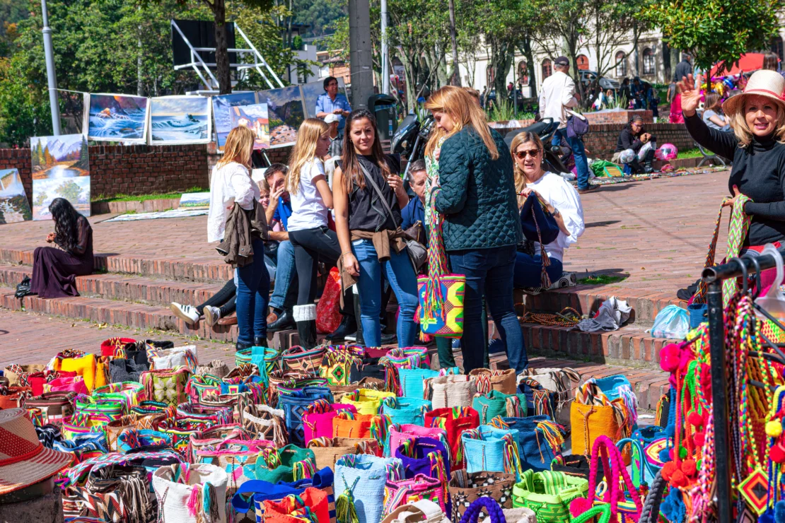 People browsing colorful bags at an outdoor market. Paintings are displayed in the background, and people are sitting and talking on steps. Trees and greenery are visible in the distance.