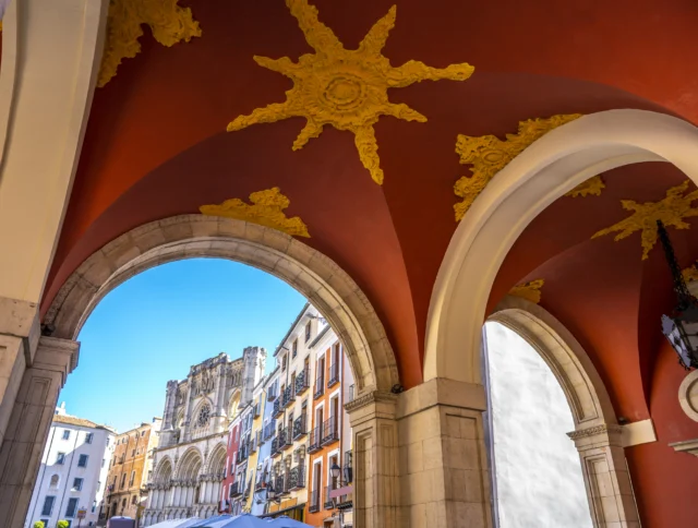 Red arched corridor with decorative yellow designs overlooks historic buildings and a cathedral in a European city under a clear blue sky.