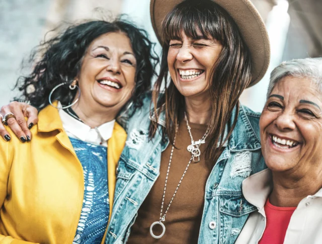 Three women are smiling and embracing each other. One wears a hat and denim jacket, another has curly hair and a yellow coat, and the third has short gray hair and a beige jacket.
