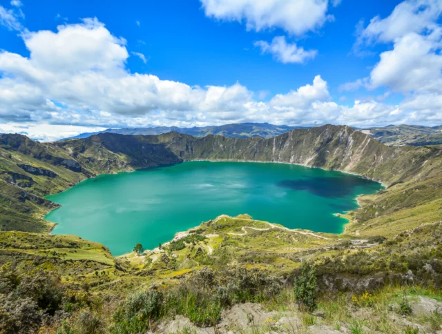 A turquoise crater lake surrounded by green hills and mountains under a partly cloudy sky.