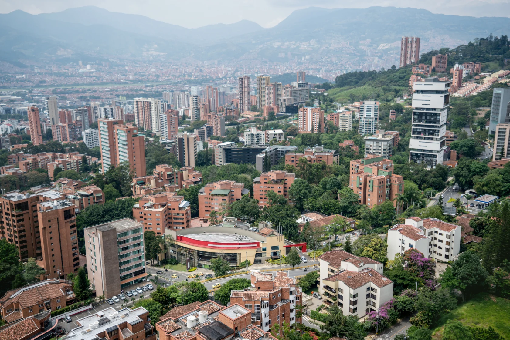 Aerial view of a sprawling cityscape with numerous brick and concrete buildings, surrounded by green hills and mountains in the background.