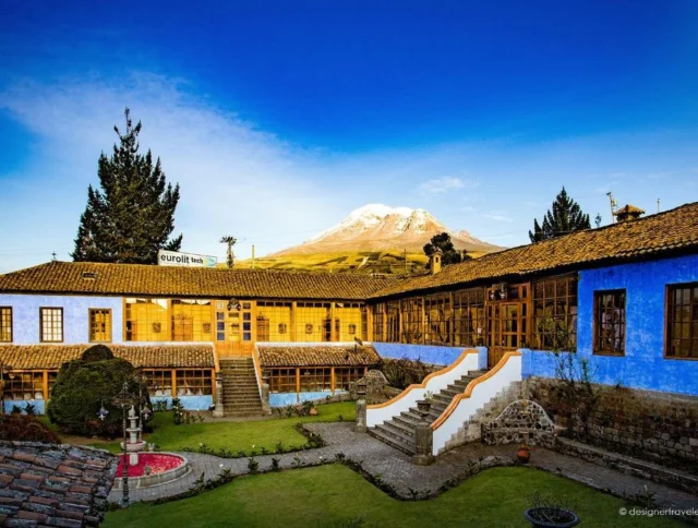 A rustic courtyard with blue and yellow buildings, surrounded by greenery, with a snow-capped mountain in the background under a clear blue sky.