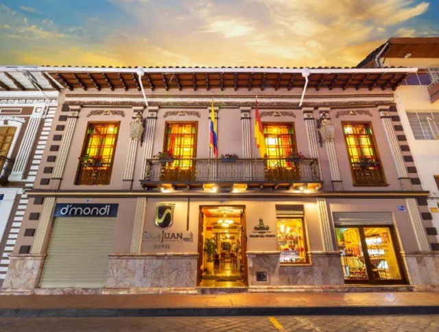 Street view of a historic building with a hotel entrance. The facade features large windows and a balcony decorated with flower boxes. A Colombian flag hangs prominently.