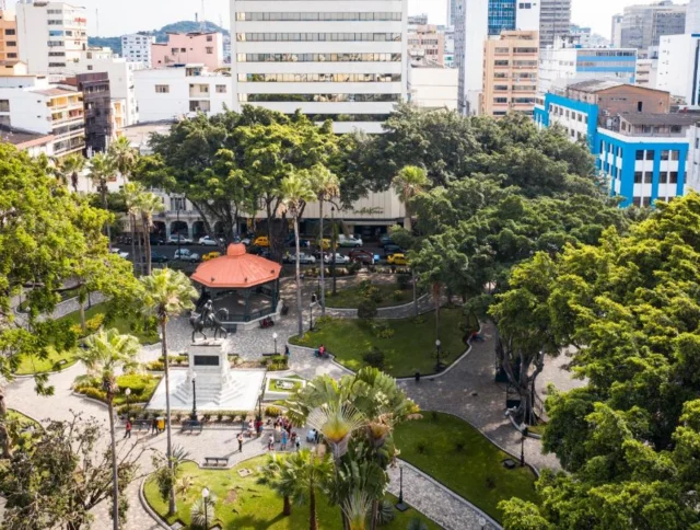 Aerial view of a city square with a central gazebo, surrounded by green trees and tall buildings in the background.