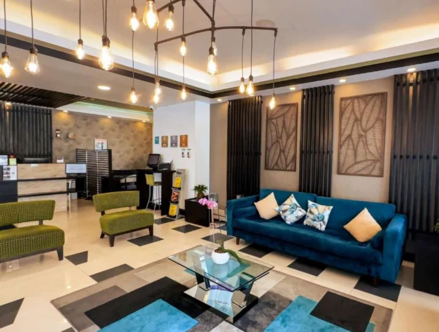 Hotel lobby with modern decor, including green armchairs, a blue sofa with pillows, a glass coffee table, and hanging light fixtures. Plants and wall art add decorative elements.