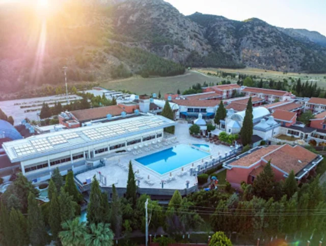 Aerial view of a resort with a central outdoor pool surrounded by red-roofed buildings, set against a backdrop of mountains and greenery.
