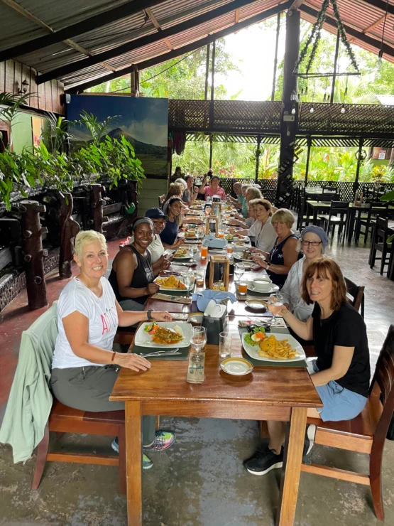 A group of people sitting at a long table in a restaurant, enjoying a meal with various dishes. The setting is open and surrounded by greenery.