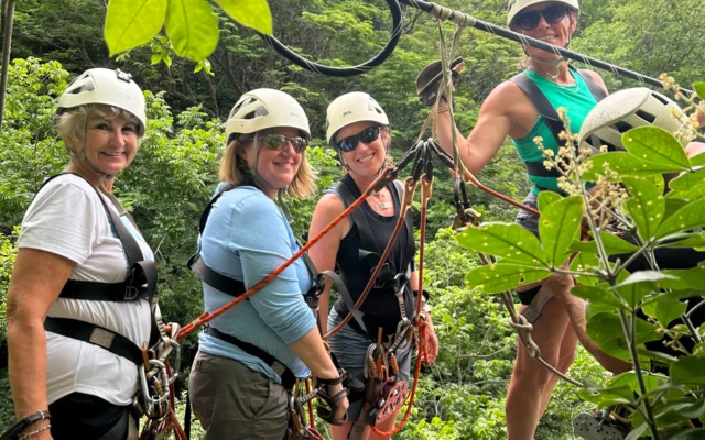 Four people in helmets and harnesses stand on a zipline platform in a lush forest setting.