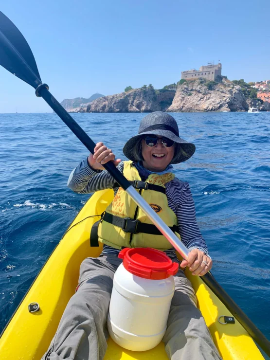 A person kayaking on the ocean, wearing a hat, sunglasses, and a life vest. There is a storage container on the kayak and a building in the background.