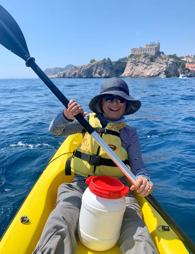 A person kayaking on the ocean, wearing a hat, sunglasses, and a life vest. There is a storage container on the kayak and a building in the background.
