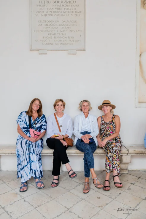 Four women sit on a bench against a white wall, smiling at the camera.