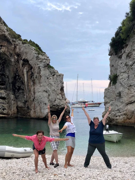 Four people pose energetically on a rocky beach with tall cliffs and anchored boats in the background.