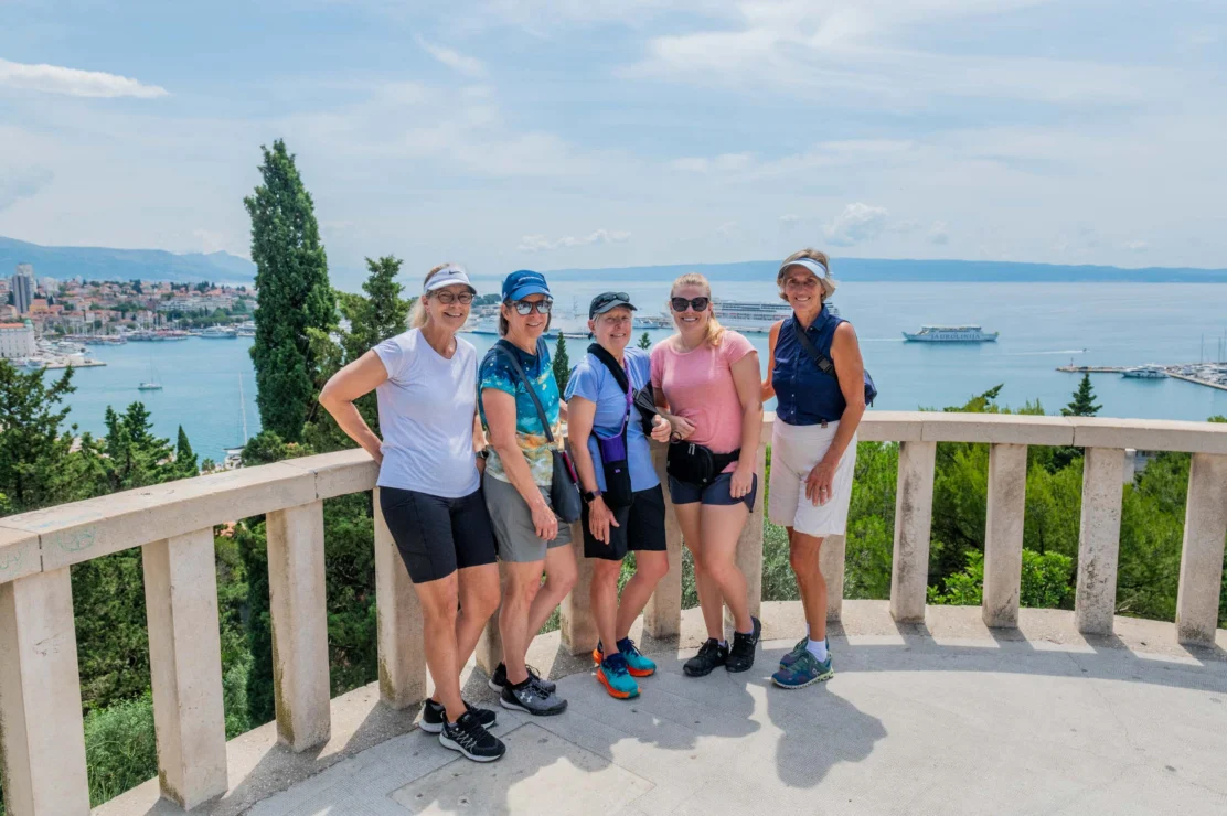 Five people stand on a balcony overlooking a coastal cityscape with a body of water and cruise ship in the background.
