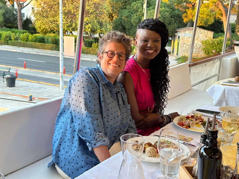 Two people sitting at an outdoor table with plates of food and glasses of wine, smiling at the camera. Trees and a street are visible in the background.