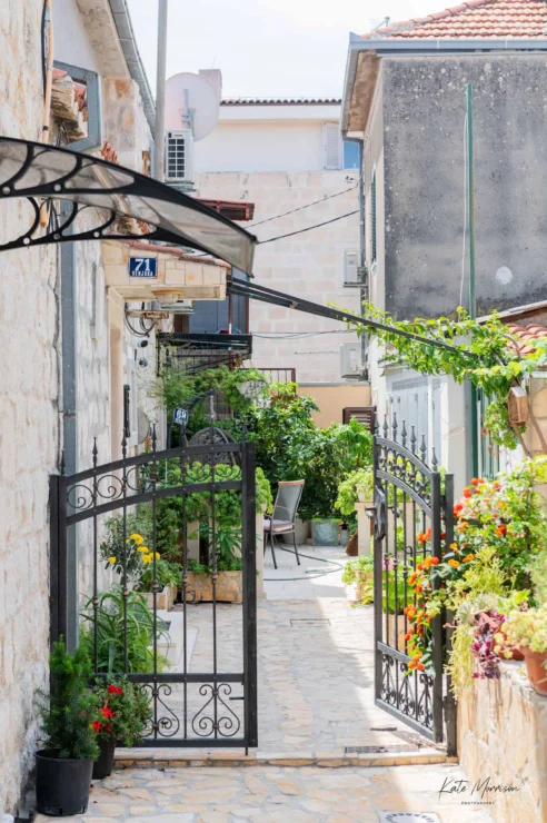 A narrow stone walkway leads through an open wrought iron gate, flanked by potted plants and flowers, towards a small courtyard with outdoor seating.