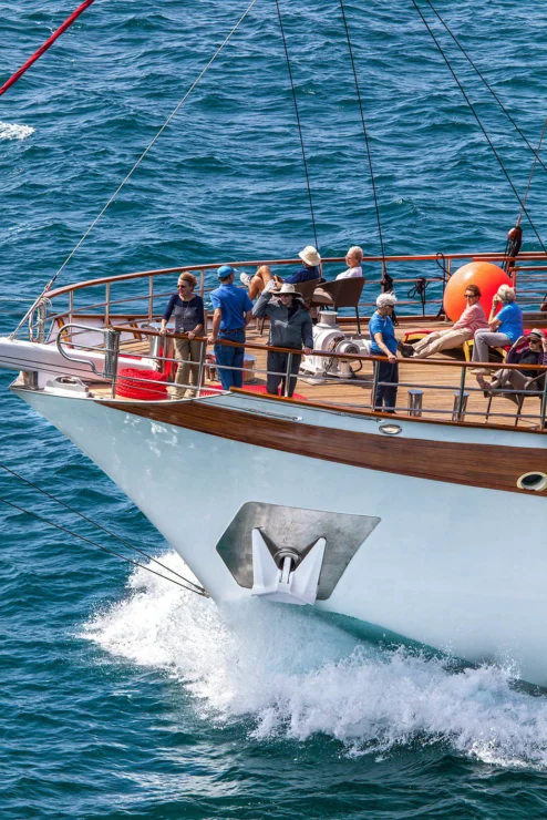 People stand and sit on the deck of a large boat sailing on a sunny day, with water splashing at the front.