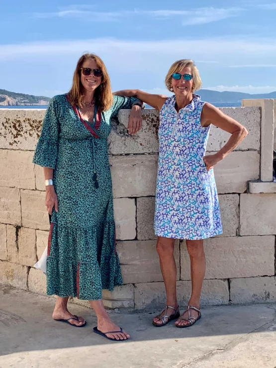 Two women in sunglasses pose next to a stone wall, with a scenic backdrop of mountains and water. One wears a long green dress, the other a blue and white dress with sandals.