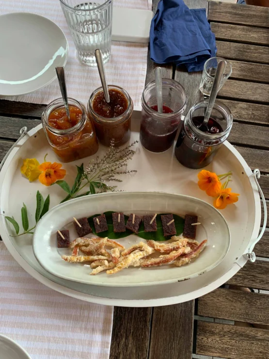 A tray with three jars of jam, a plate of sliced desserts, orange flowers, and a glass on a wooden table.