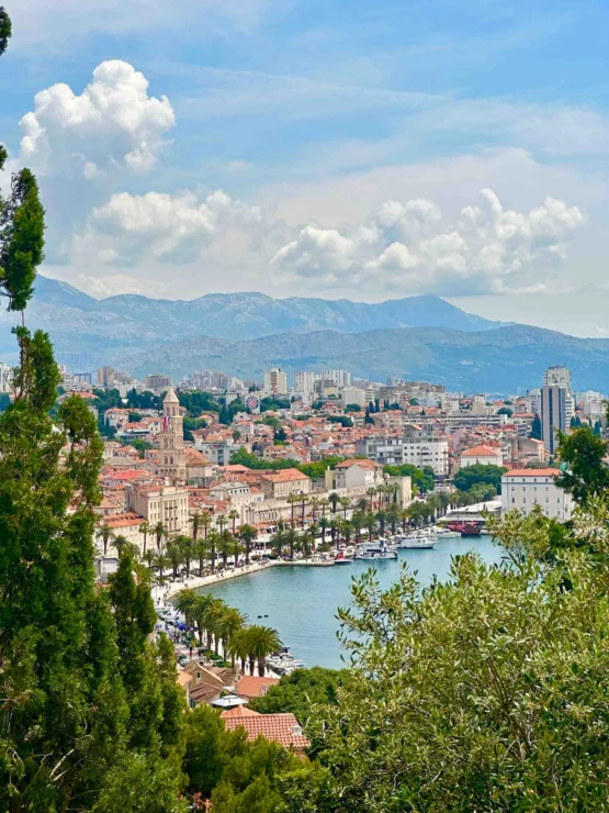 View of a coastal city with red-roofed buildings, a harbor lined with palm trees, and mountains in the background under a partly cloudy sky.