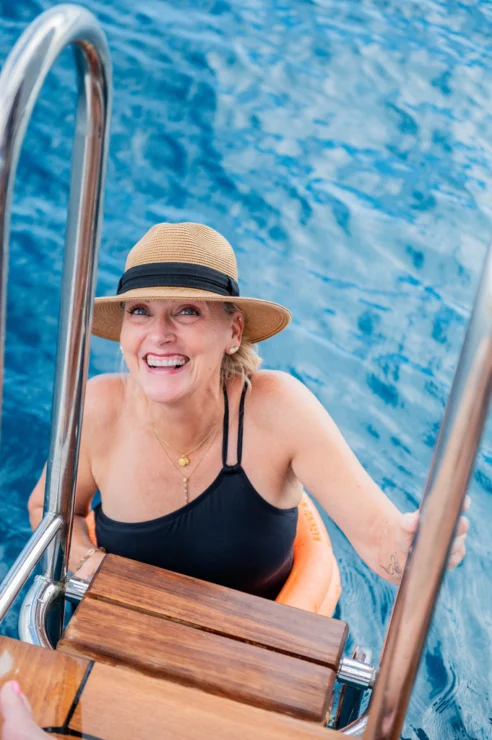 A woman in a black swimsuit and straw hat smiles while holding onto a metal ladder in a pool or body of water.
