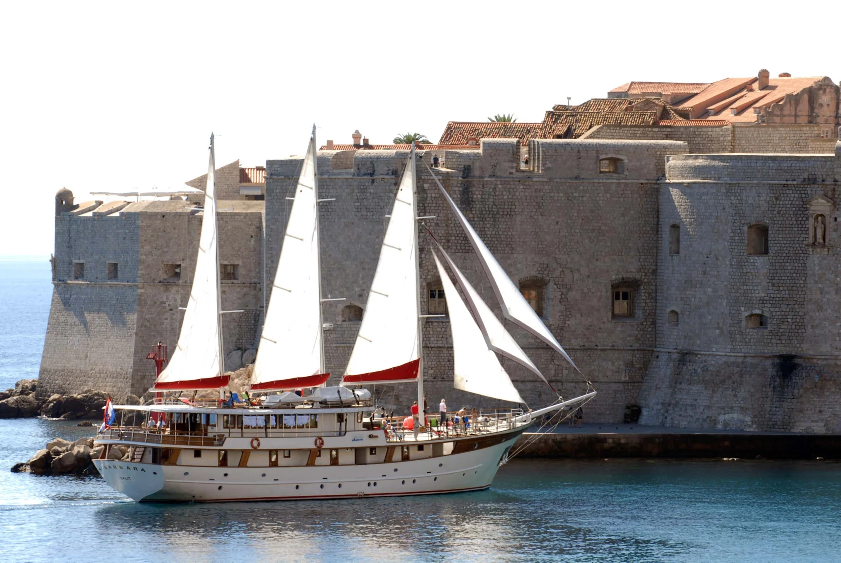 A sailboat with three sails glides near an ancient stone fortress by the sea, under a clear sky.