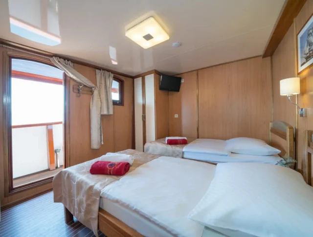 A tidy cabin room with two single beds, white linens, and red towels. There's a wall-mounted TV, a window with curtains, and an open door leading to a small balcony.