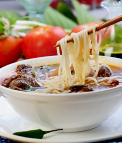 A bowl of noodle soup with meat and vegetables, with chopsticks lifting noodles. Fresh tomatoes and greens in the background.