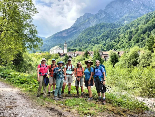 A group of people in hiking gear stand on a dirt path with a mountain village and lush greenery in the background.