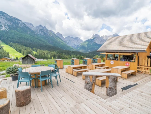 Outdoor seating area with wooden tables and chairs on a deck. Mountains and green fields in the background under a partly cloudy sky.