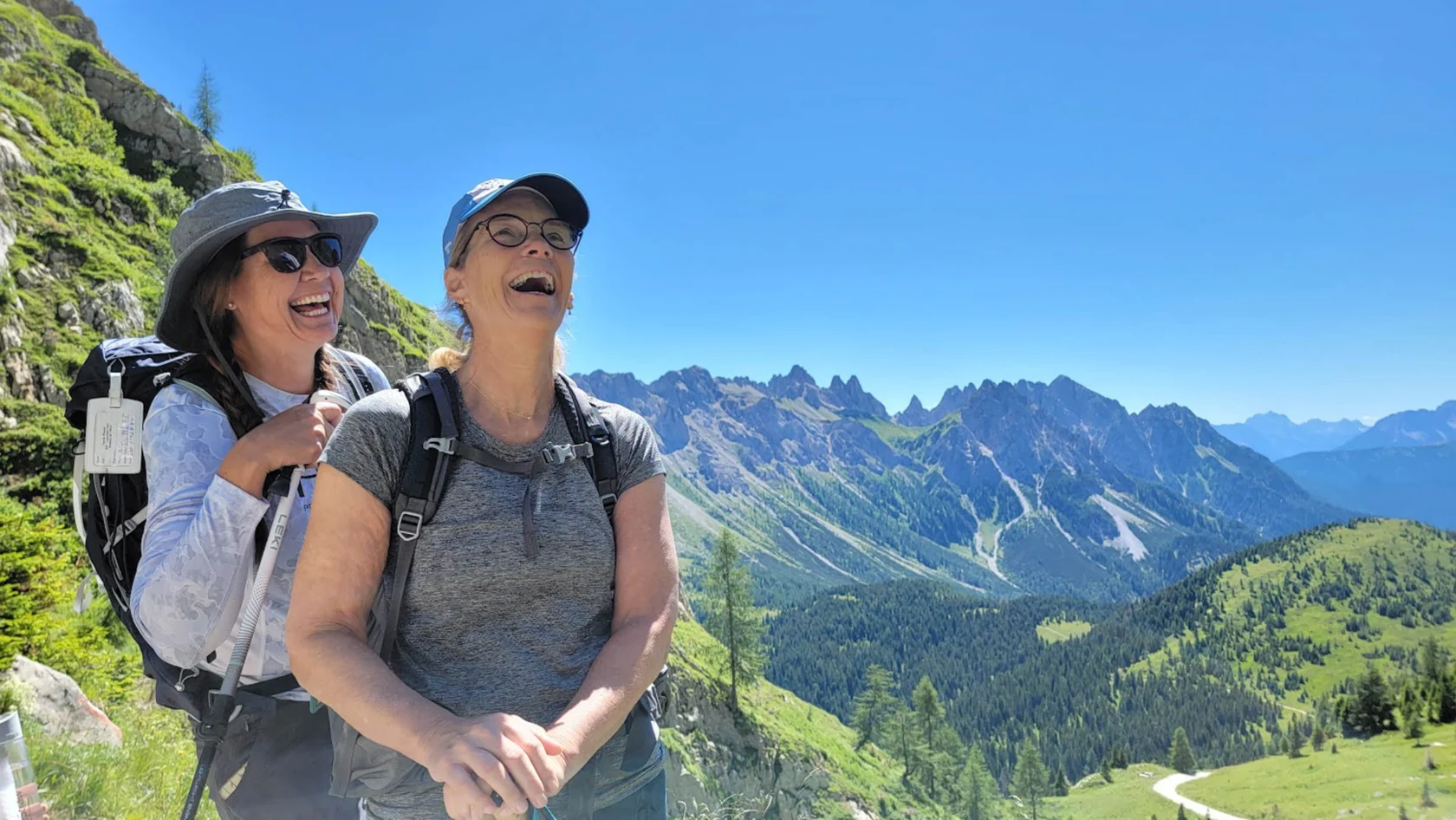 Two women in hiking gear laugh together on a mountain trail with a scenic backdrop of peaks and blue sky.