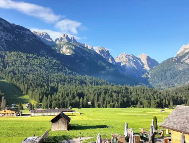 A scenic view of a green valley with wooden buildings, surrounded by dense forests and rugged mountain peaks under a clear blue sky.