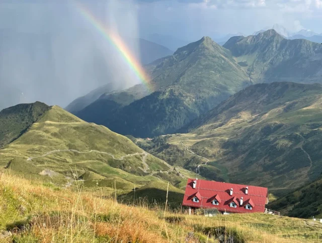 Mountain landscape with a red-roofed building in the foreground and a rainbow stretching across the sky. The scene includes green hills and distant peaks.