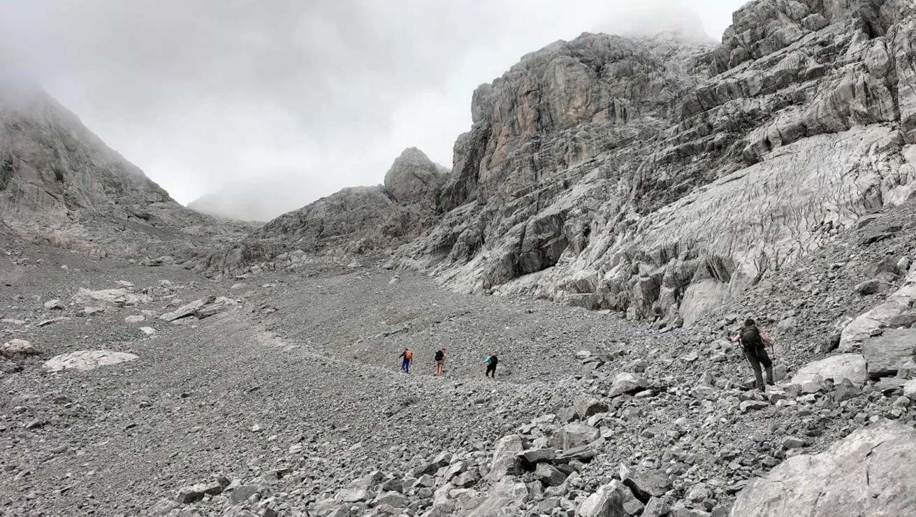 Hikers traverse a rocky, gray mountainous landscape under a cloudy sky.