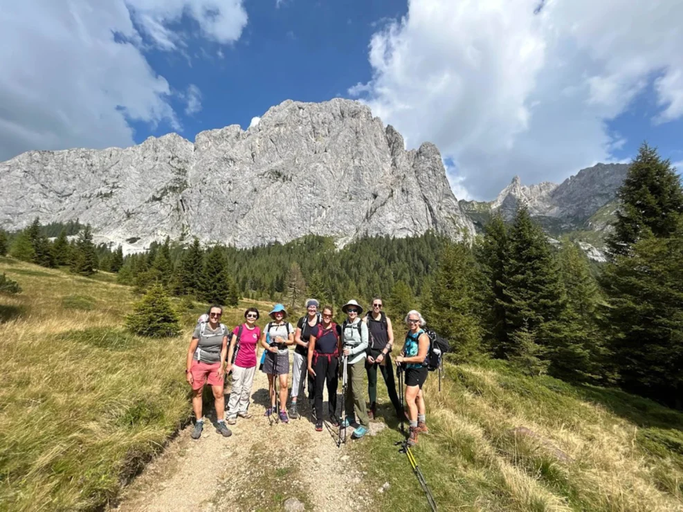 A group of hikers stands on a trail in a mountainous landscape with rocky peaks and green trees under a partly cloudy sky.