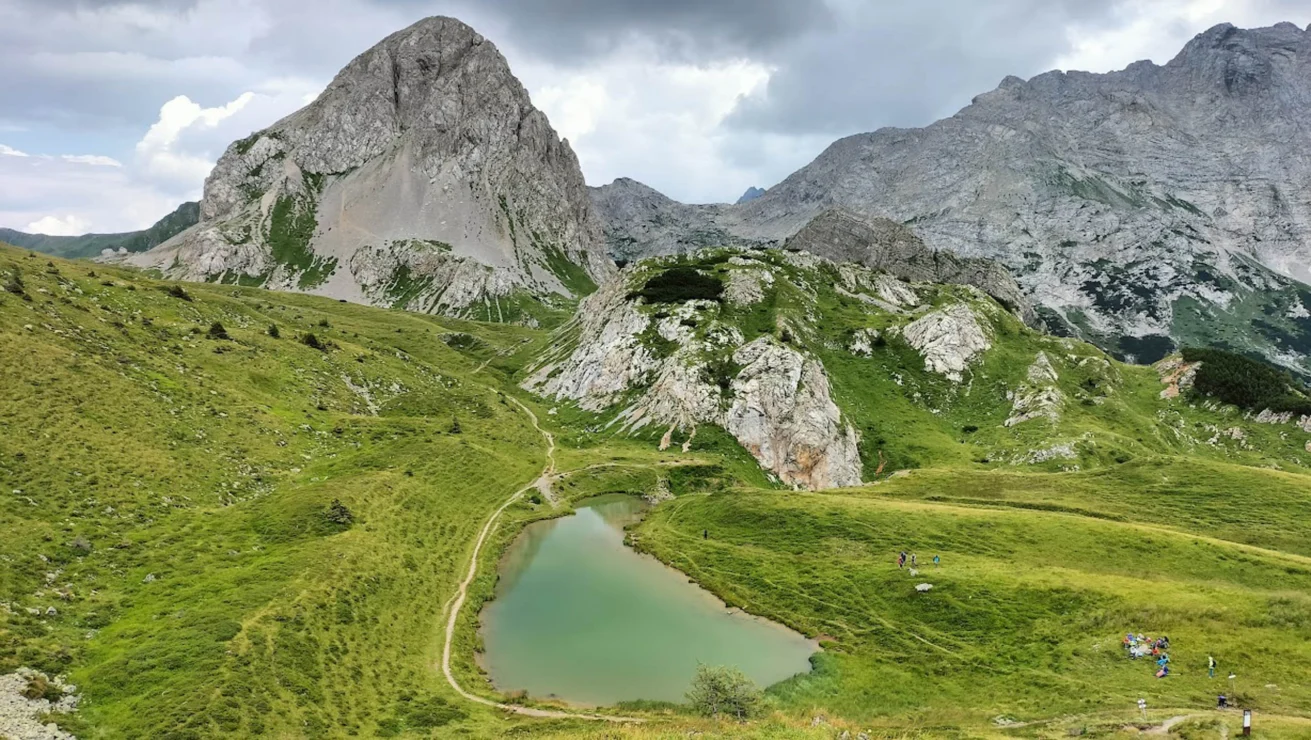 Mountain landscape with a small lake surrounded by grassy hills and rocky peaks under a cloudy sky. Several people are visible near the water.