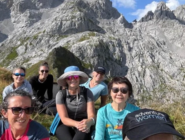 A group of people sitting on grass in front of rocky mountains under a blue sky.