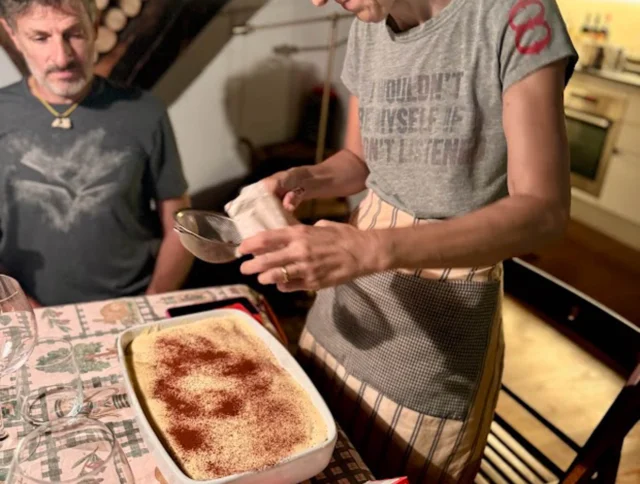 A woman sprinkles cocoa powder on a dish while a man sits at the table. The scene is set in a cozy kitchen with wood details.