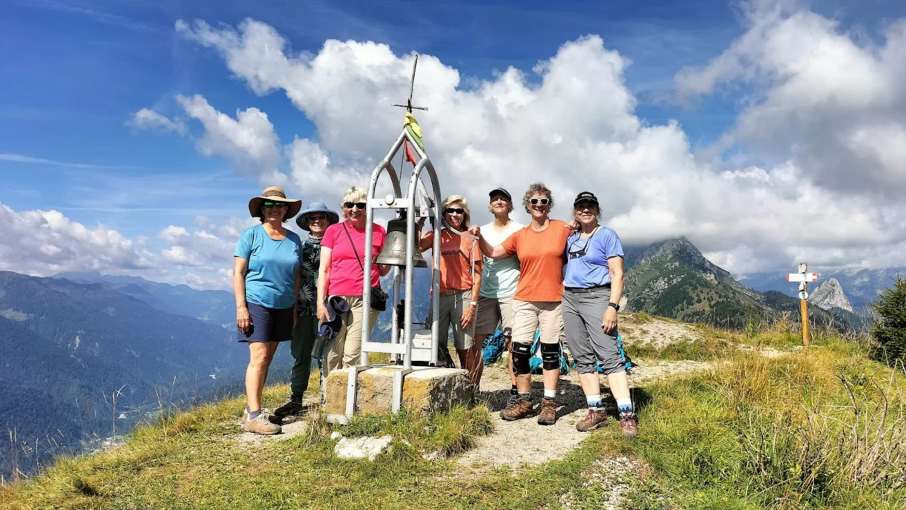 A group of people standing on a mountain summit next to a small cross and a plaque, with a scenic view of mountains and clouds in the background.