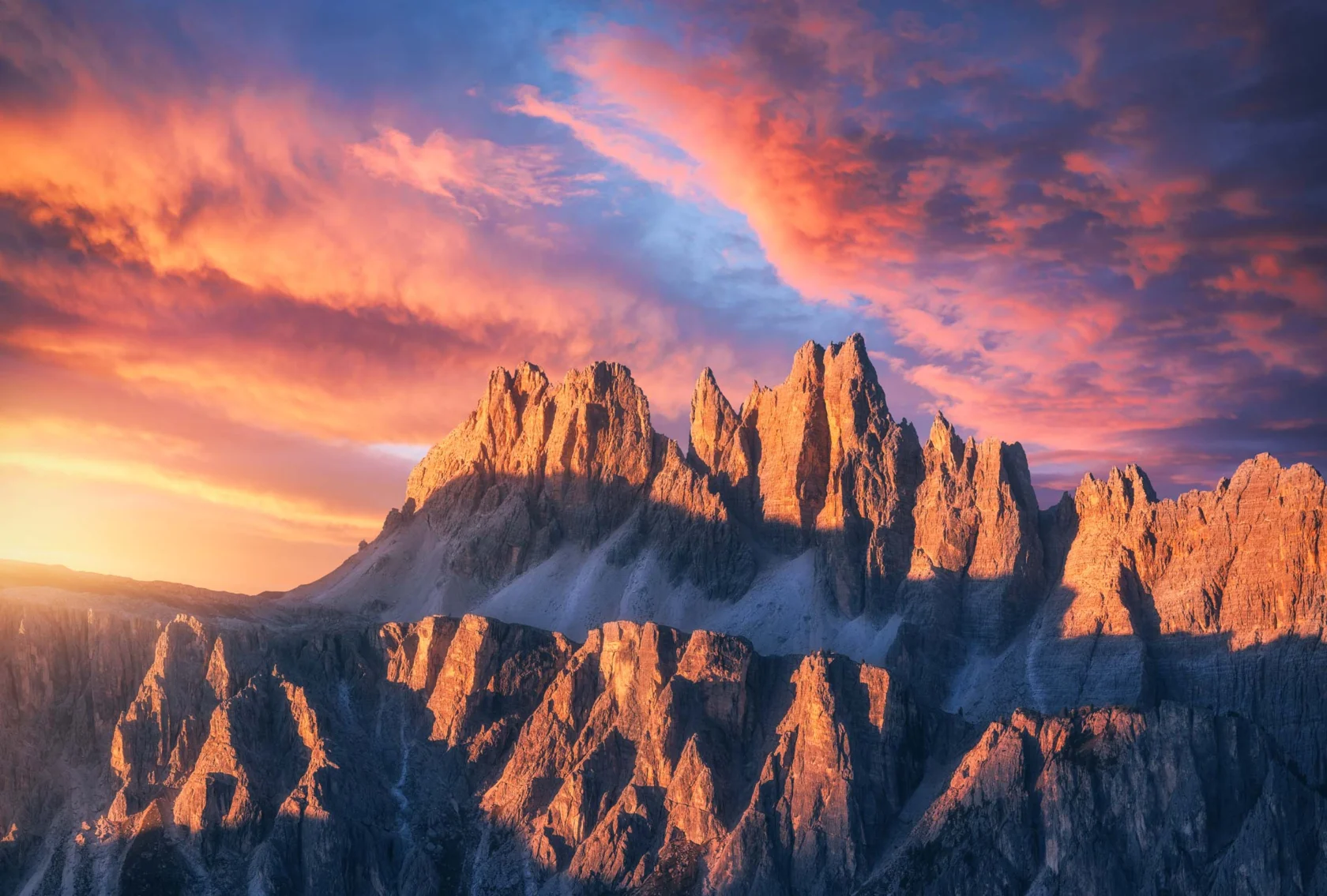 A mountain range with jagged peaks is bathed in orange and pink light from a vibrant sunset sky.