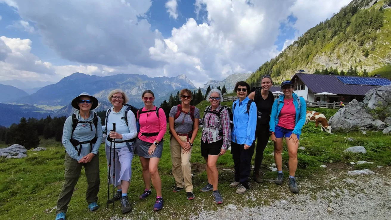A group of eight women posing for a photo in a mountainous outdoor setting. They are dressed in hiking attire, with a scenic view of mountains and a cabin in the background.