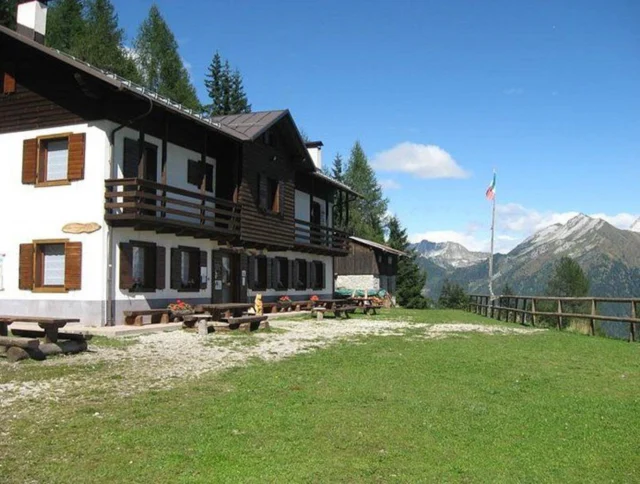 A rustic wooden lodge with a balcony stands against a backdrop of mountains and trees under a blue sky. An Italian flag is seen on a flagpole nearby.