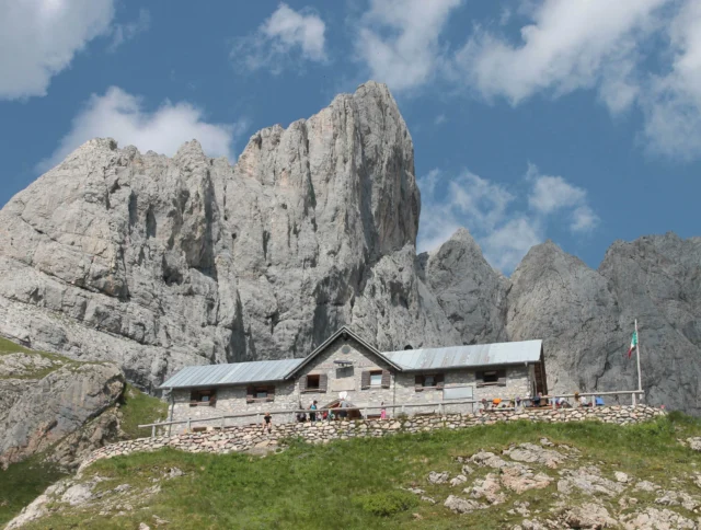 A rustic stone building with a metal roof sits in front of towering rocky mountains under a partly cloudy sky. A flag is visible on the right.