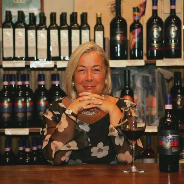 A woman with short hair sits at a table with a glass and bottle of red wine. Shelves lined with wine bottles fill the background.