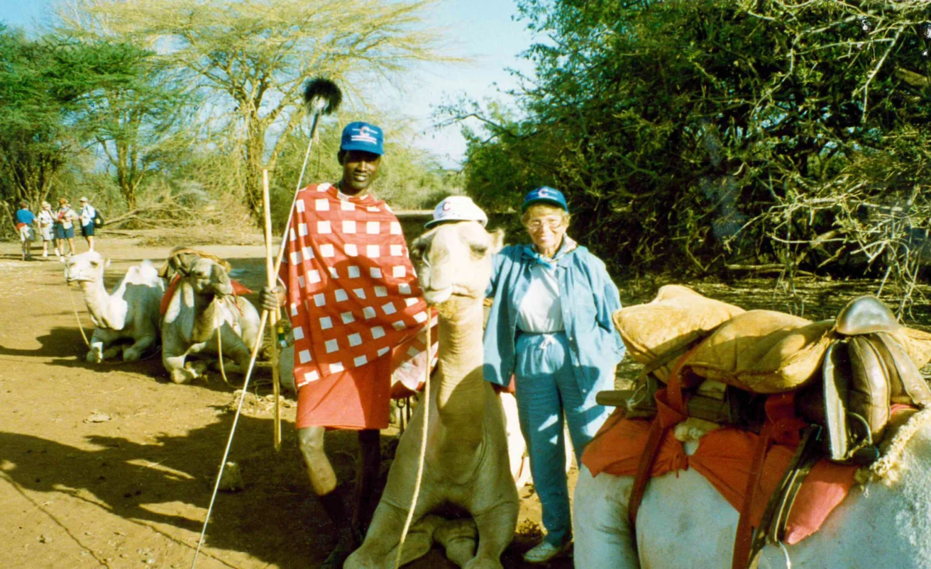 A person in traditional clothing and a person in casual attire stand with camels wearing saddles and hats in a rural area with trees in the background.
