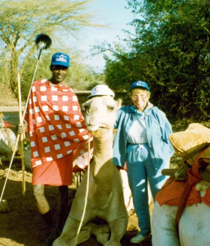 A person in traditional clothing and a person in casual attire stand with camels wearing saddles and hats in a rural area with trees in the background.
