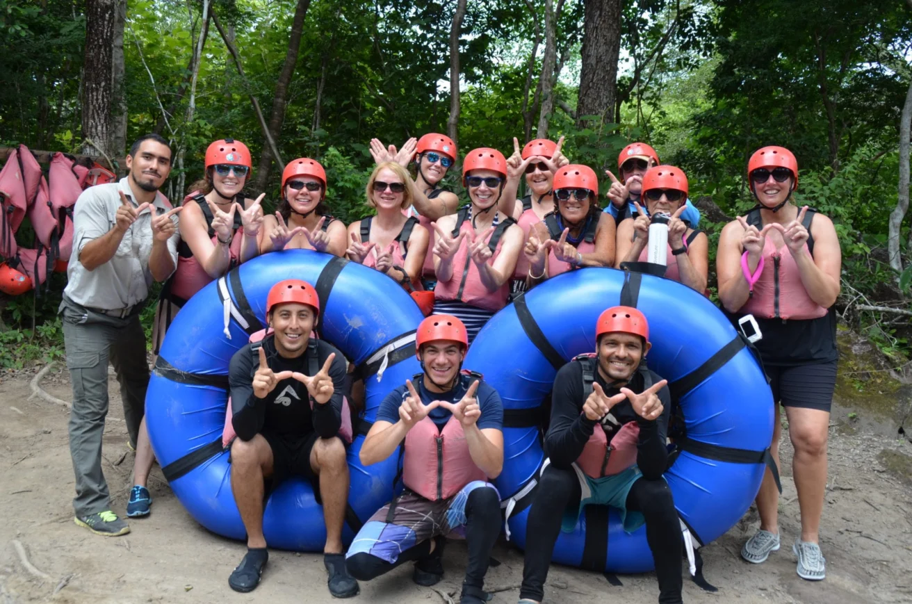Group of people wearing helmets and life jackets posing with blue rafts in a forested area.
