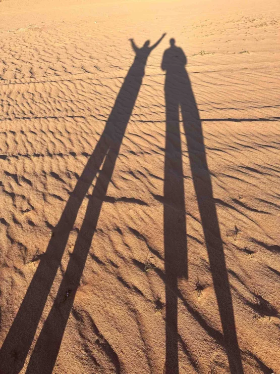 Two long shadows on sandy terrain, one with raised arms.