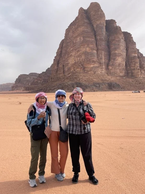 Three people in casual clothing stand together in front of a large rock formation in a desert landscape.